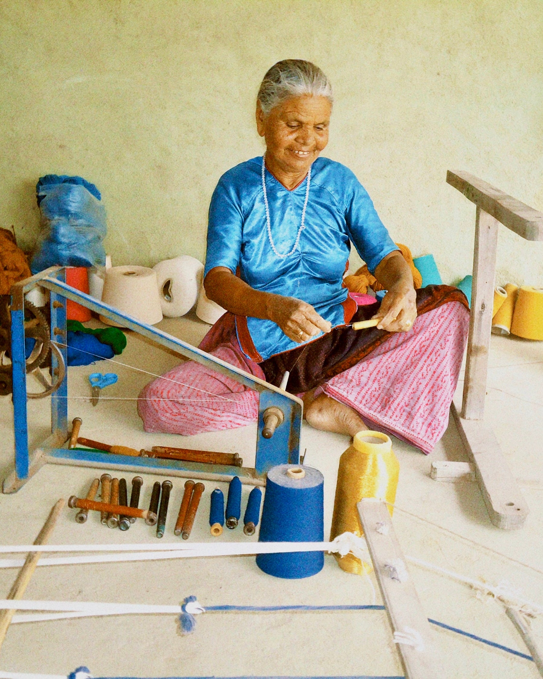 An elderly woman sits cross-legged on a floor surrounded by textile tools and spools of thread, wearing a bright blue top and pink patterned skirt while working with fabric.