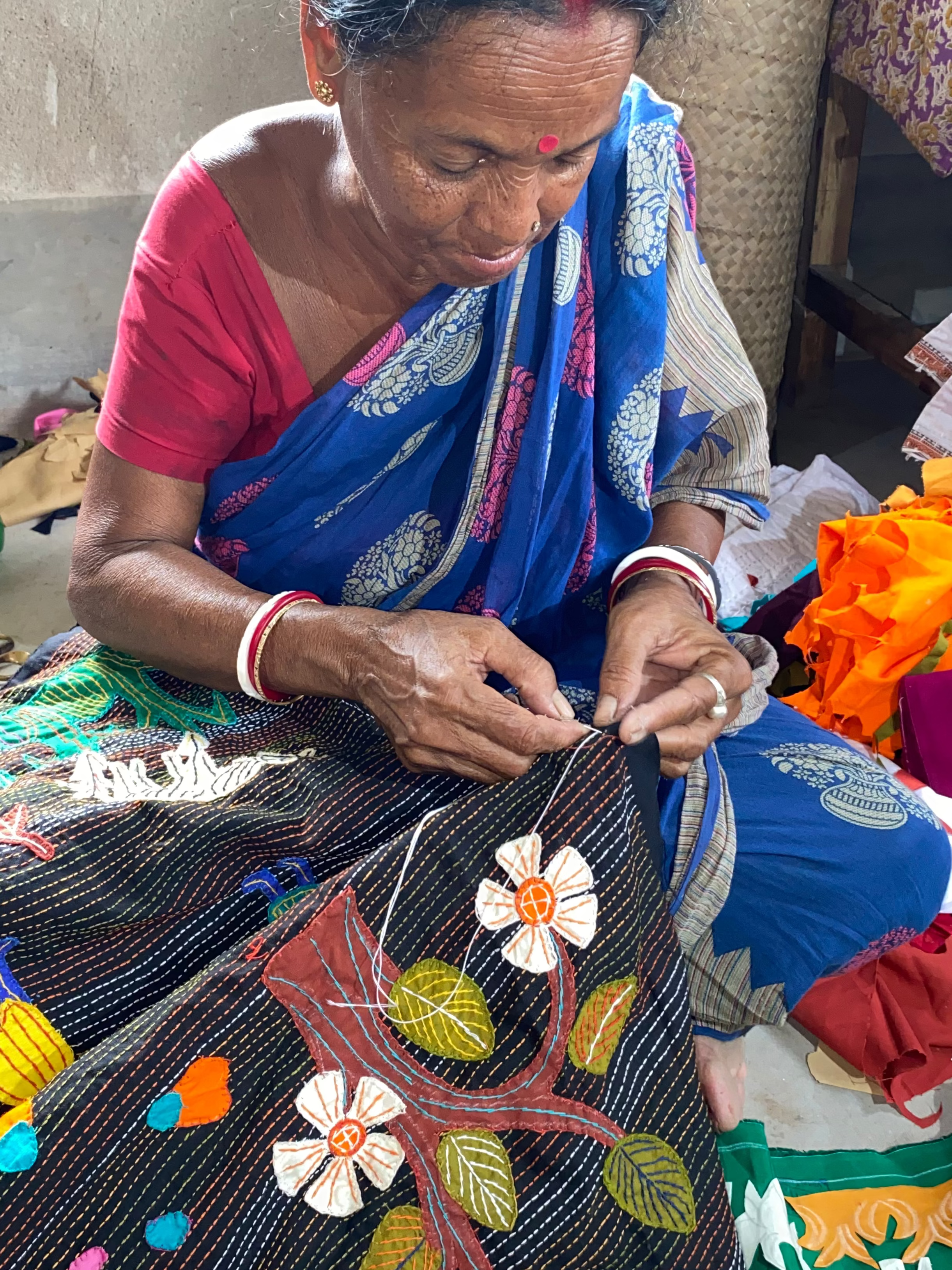 An elderly woman in a blue and pink sari is seated on the floor, hand-stitching a black embroidered fabric piece decorated with white flowers and green leaves. The background shows a rustic indoor setting with other colorful textiles and a green plastic bottle.