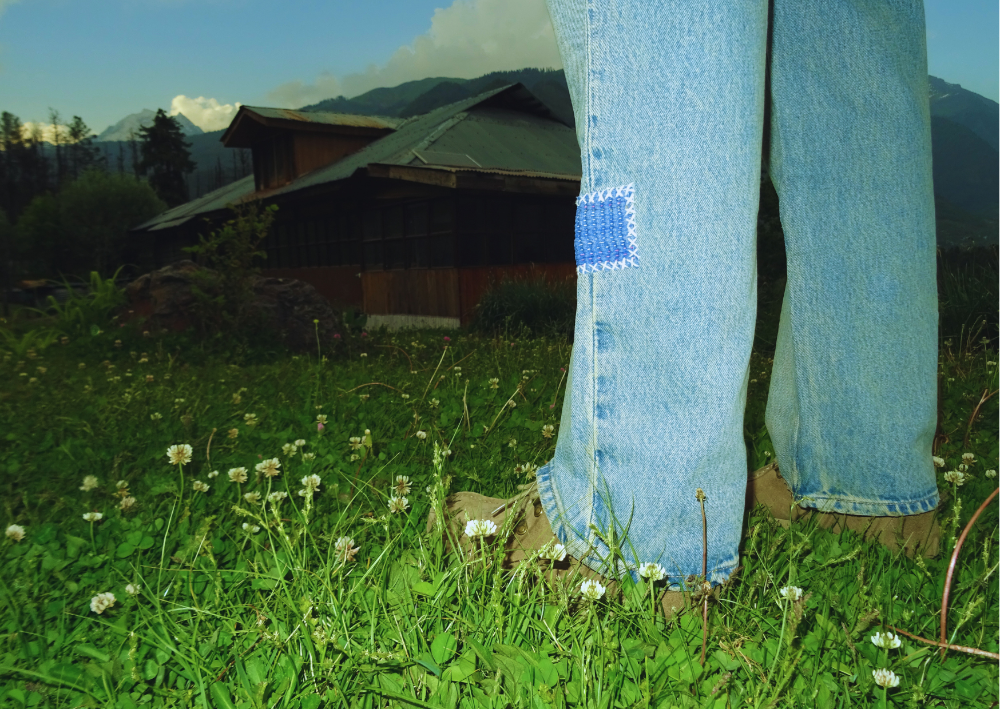 Person wearing blue jeans with a patch on a grassy field with a cabin in the background