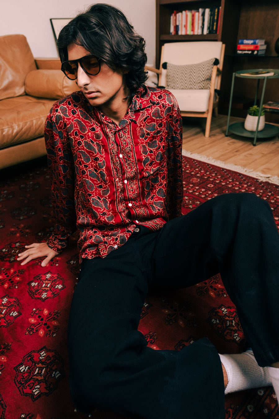 Person wearing a red patterned shirt sitting on a red rug in a living room.