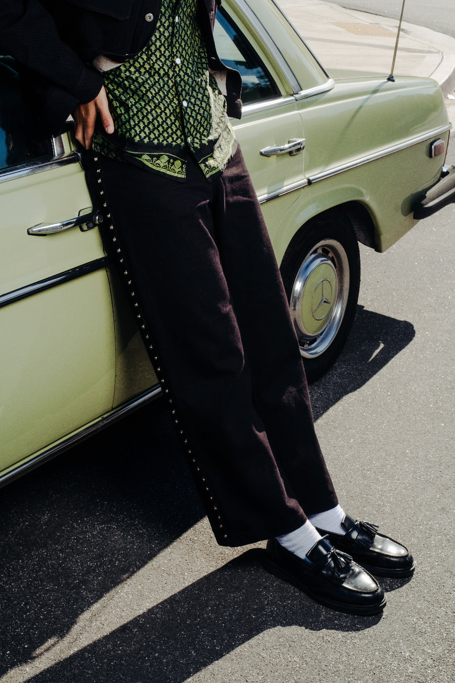 Person wearing a patterned shirt, dark pants, and black shoes standing next to a vintage car.