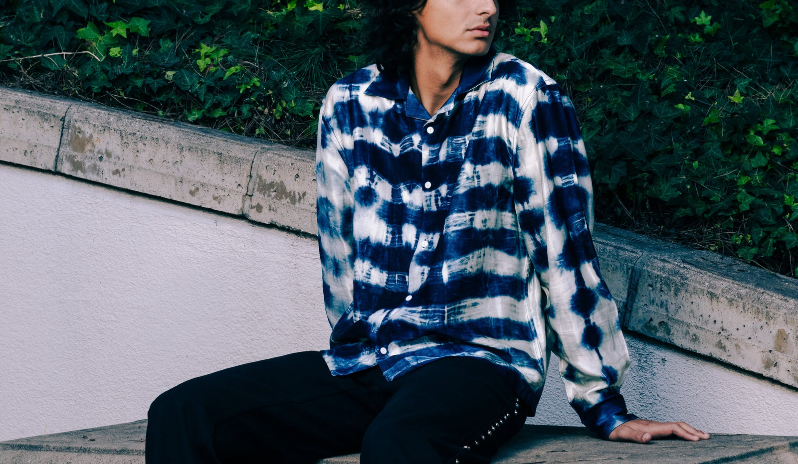 Person wearing a blue and white checkered shirt sitting on a concrete bench with greenery in the background