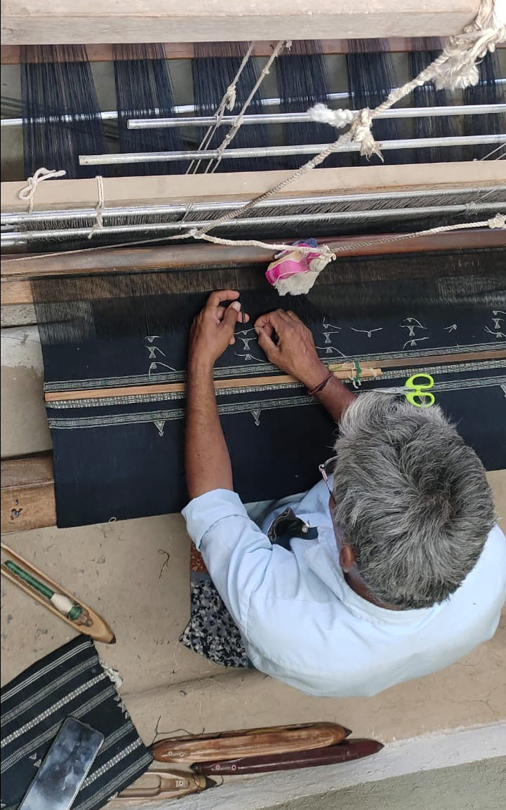 A person with gray hair works at a traditional handloom, weaving a striped textile. The scene shows wooden loom components, threads, and the artisan's hands adjusting the weaving process.