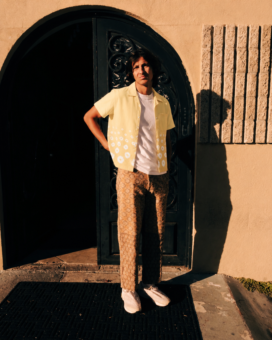 Person wearing a yellow floral-print shirt and matching trousers in front of an arched doorway – Hindostan Archive